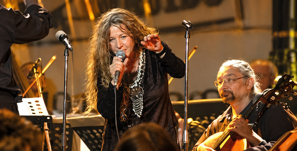 Claudia Puyó canta con un micrófono durante una actuación, acompañada por músicos en el escenario.