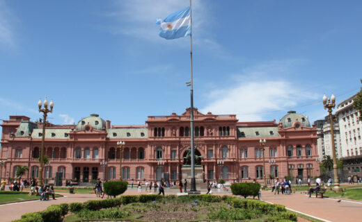 Frente completo de la Casa Rosada visto desde la Plaza de Mayo, con su arco central, la bandera argentina y personas caminando por la plaza