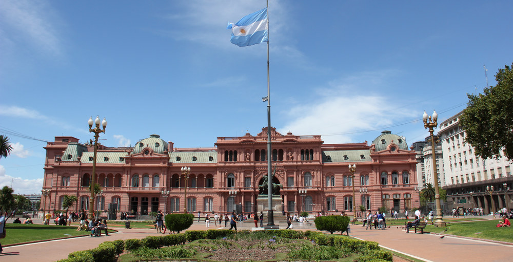 Frente completo de la Casa Rosada visto desde la Plaza de Mayo, con su arco central, la bandera argentina y personas caminando por la plaza