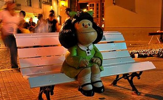 Escultura de Mafalda sentada en un banco blanco en San Telmo, con personas y edificios del barrio al fondo.