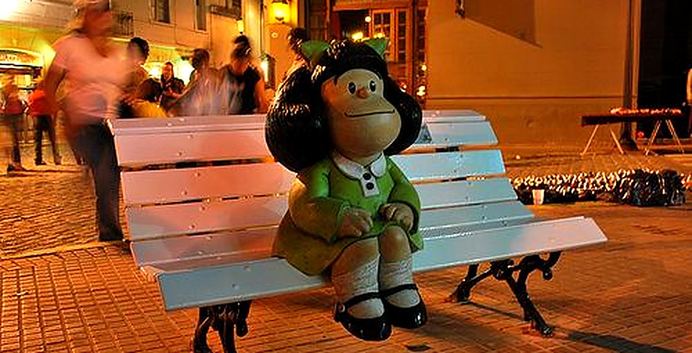 Escultura de Mafalda sentada en un banco blanco en San Telmo, con personas y edificios del barrio al fondo.