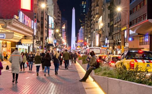 Avenida Corrientes de noche, con peatones, marquesinas iluminadas y el Obelisco al fondo, en una nota sobre sus 90 años como avenida y su historia cultural porteña.