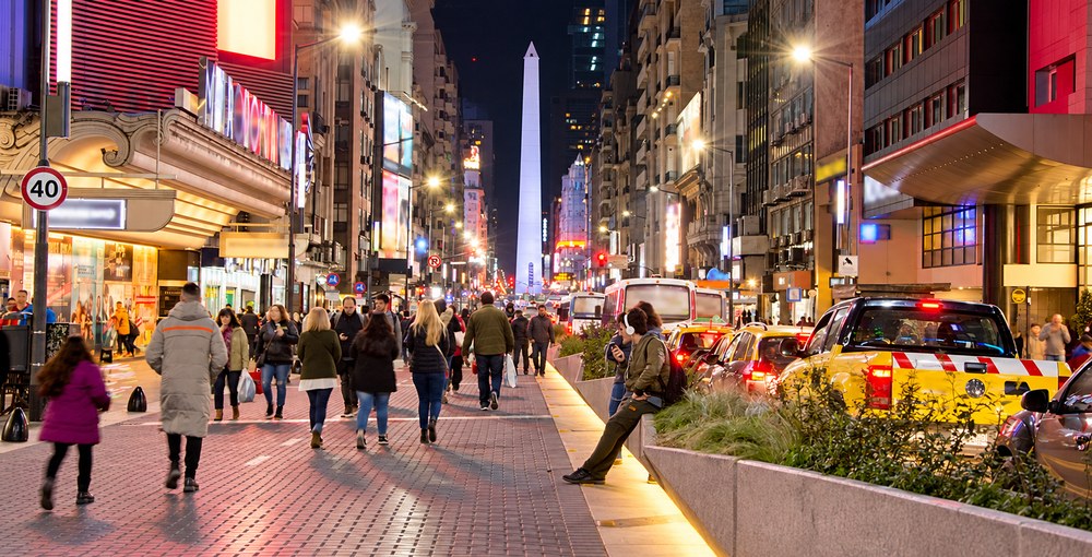 Avenida Corrientes de noche, con peatones, marquesinas iluminadas y el Obelisco al fondo, en una nota sobre sus 90 años como avenida y su historia cultural porteña.