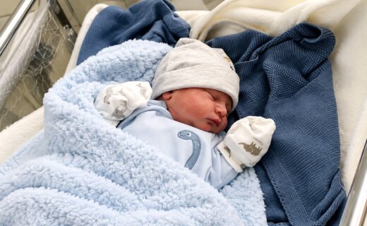Bebé recién nacido dormido en una cuna, con gorrito gris y mantas azules, para una nota sobre nombres de varones.