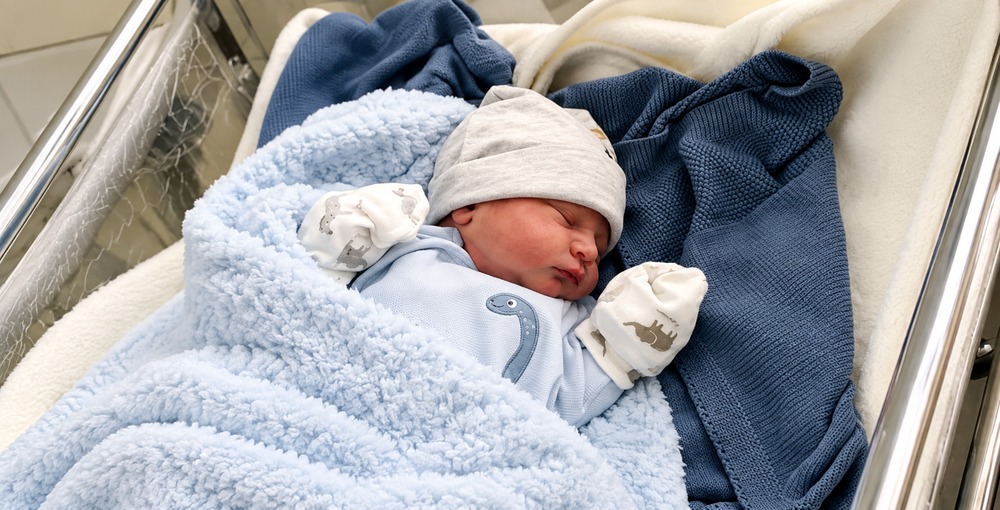 Bebé recién nacido dormido en una cuna, con gorrito gris y mantas azules, para una nota sobre nombres de varones.